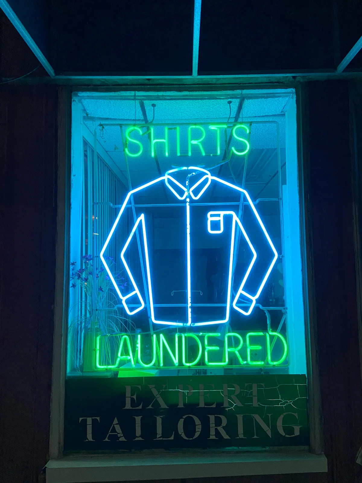Neonskylt med skjorta och texten 'Shirts Laundered', syns i ett skyltfönster på natten.