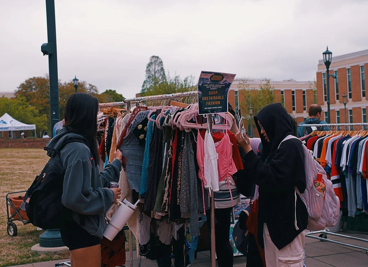 Två personer bläddrar bland second hand-kläder på marknad, skylt med 'Shop Sustainable Fashion', utomhus.