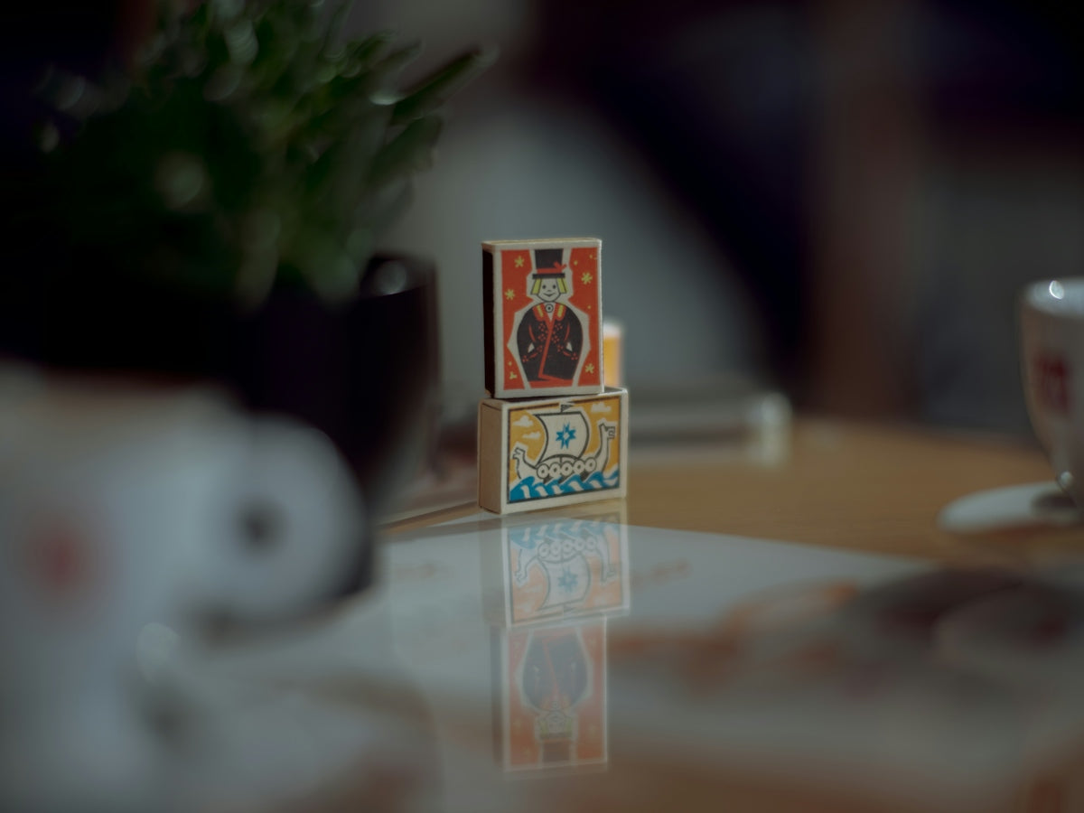 Two stacked matchboxes with illustrated characters on a reflective surface
