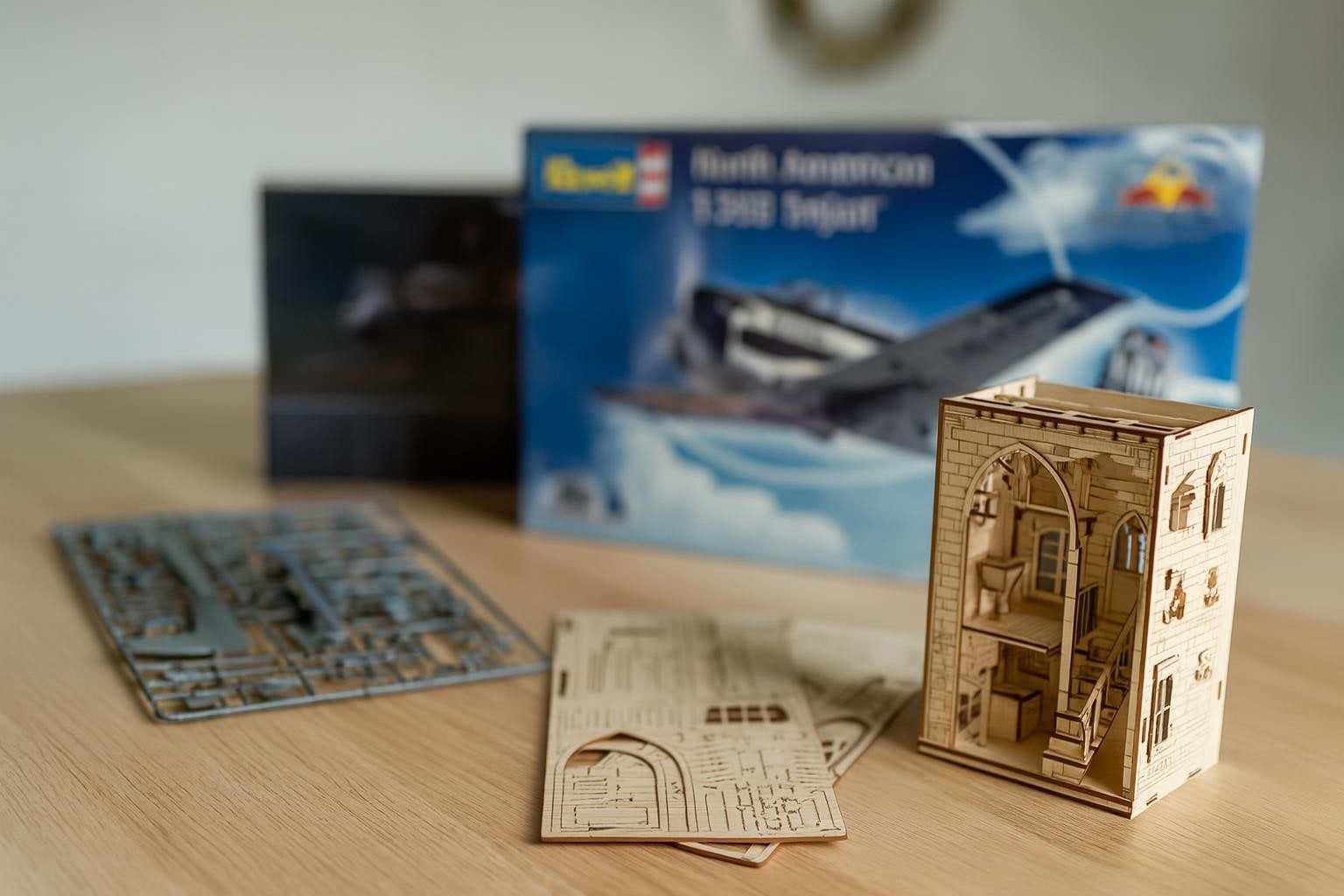 Fotorealistisk bild av modellbyggsatser på ett hobbybord med plastdelar, träbyggsats och halvfärdig book nook i mjukt dagsljus.